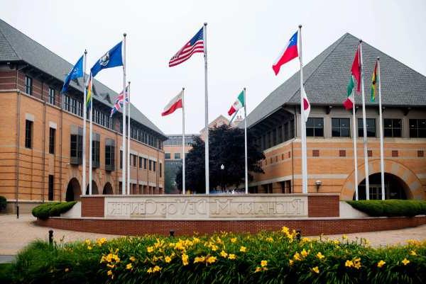 Several flags on flagpoles fly outside of a building. A cement sign says "Richard M. DeVos Center."